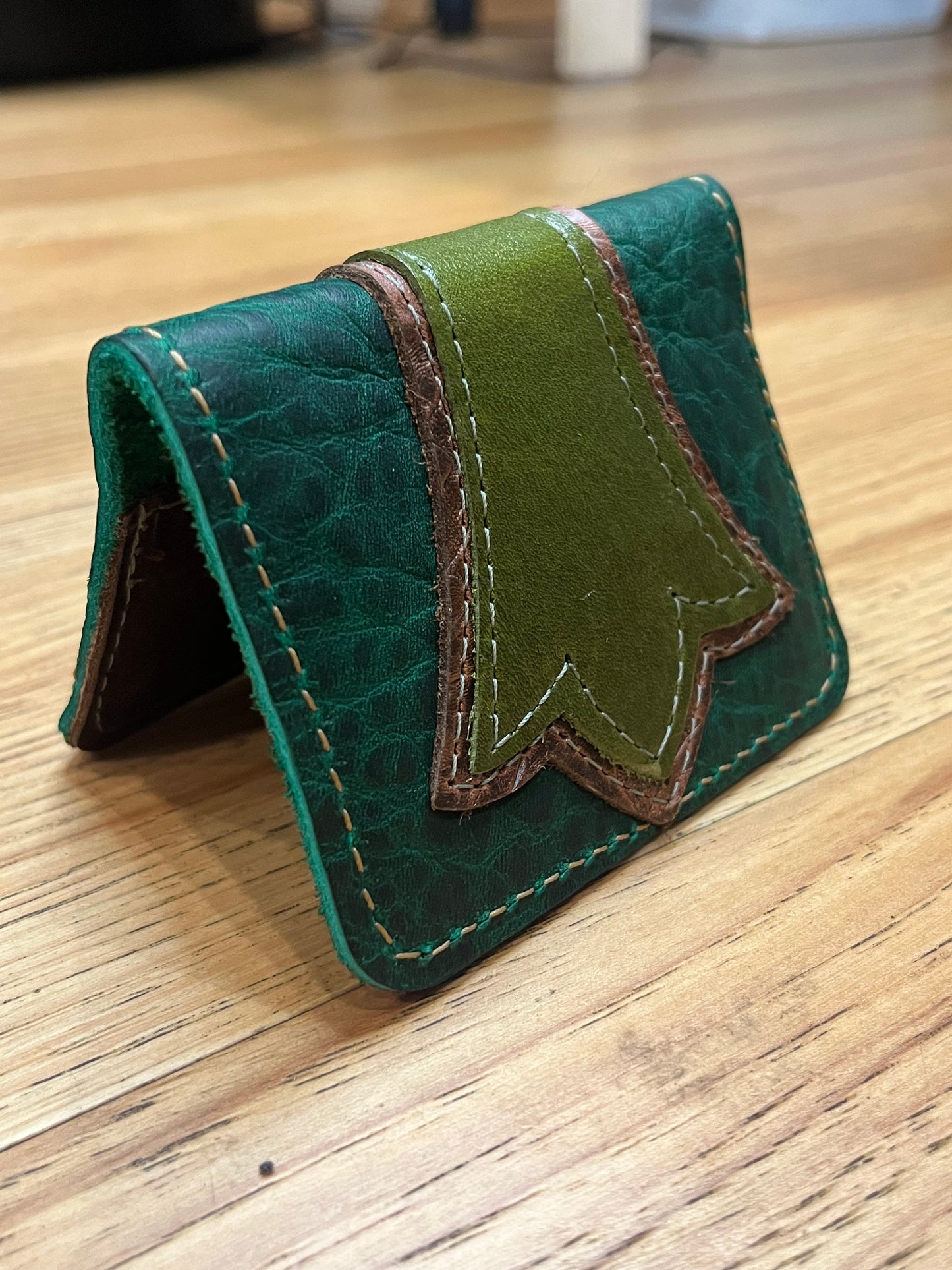 A wallet folded in half showing a base color of dark green leather with a layered trident accent in brown floral and light green leather with green and brown stitching.
