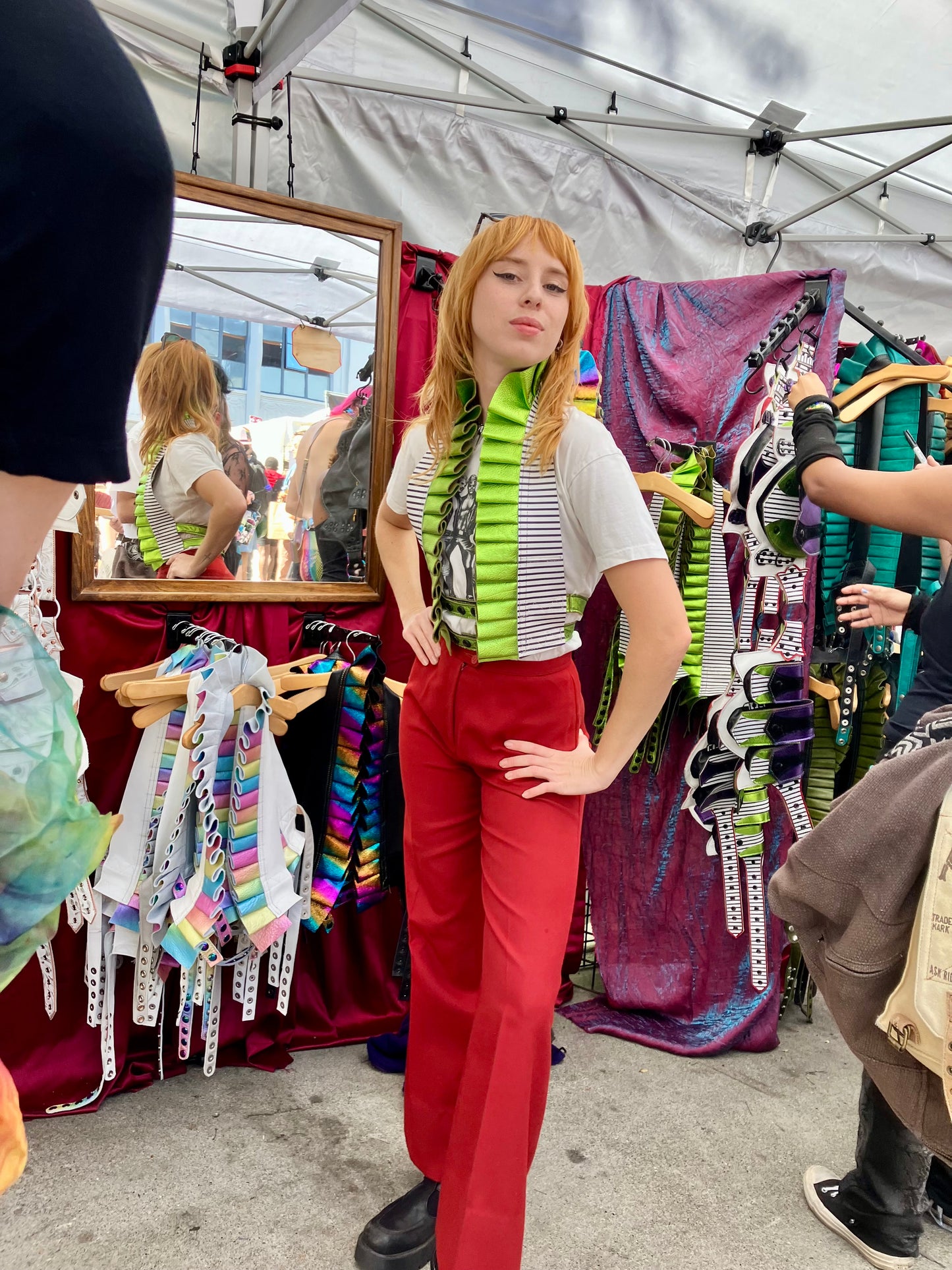 femme caucasian individual in a graphic tshirt and red pants standing infront of a small mirror and the folsom street fair booth, hands on hips looking at the viewer while sporting a showtime ruff top