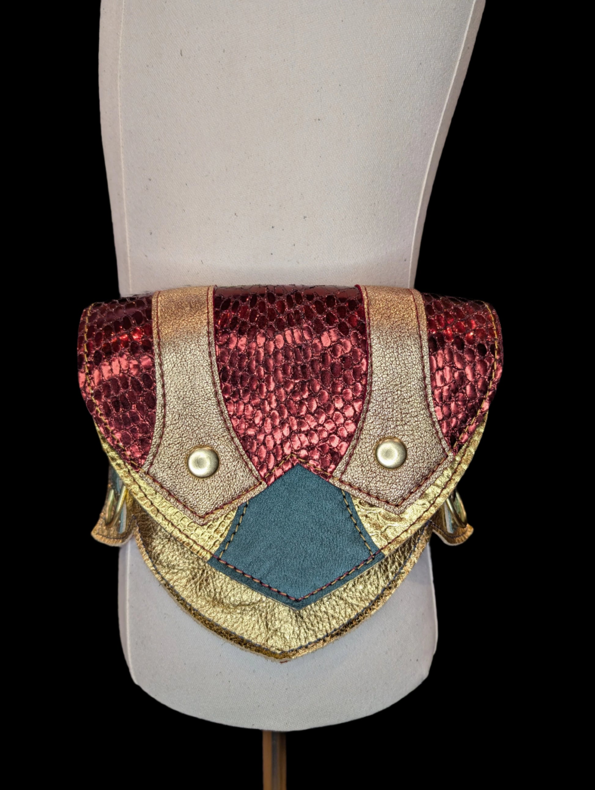 A regal belt on a mannequin with one large pocket and 3 smaller pockets made with a gold snake patterned leather with  dark green accent and flaps made with a red snake patterned leather with a gold leather accent. Pockets have a blue gusset and gold hardware.