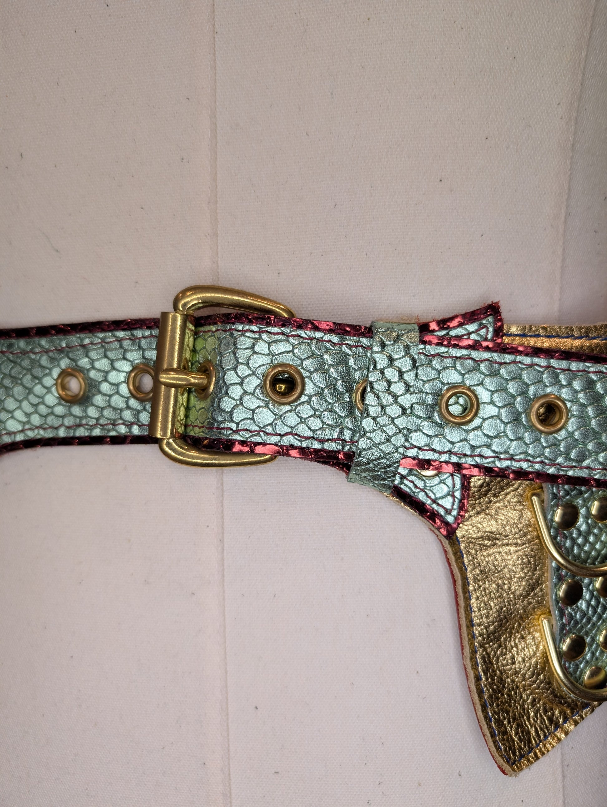 An up close photo of the buckle on a regal belt on a mannequin with a red snake patterned leather layered under a light green snake patterned leather with gold hardware.