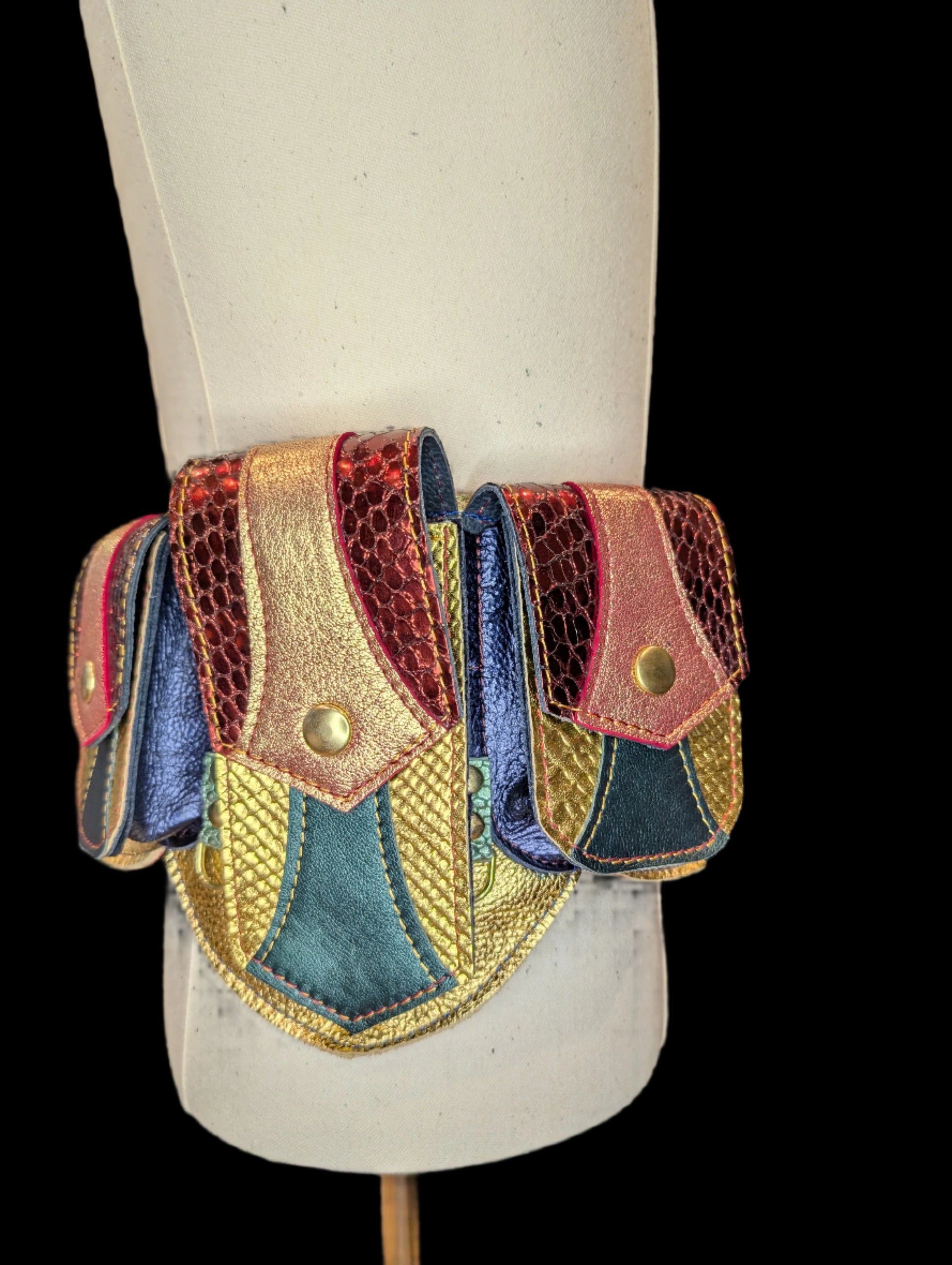 A regal belt on a mannequin with one large pocket and 3 smaller pockets made with a gold snake patterned leather with  dark green accent and flaps made with a red snake patterned leather with a gold leather accent. Pockets have a blue gusset and gold hardware.