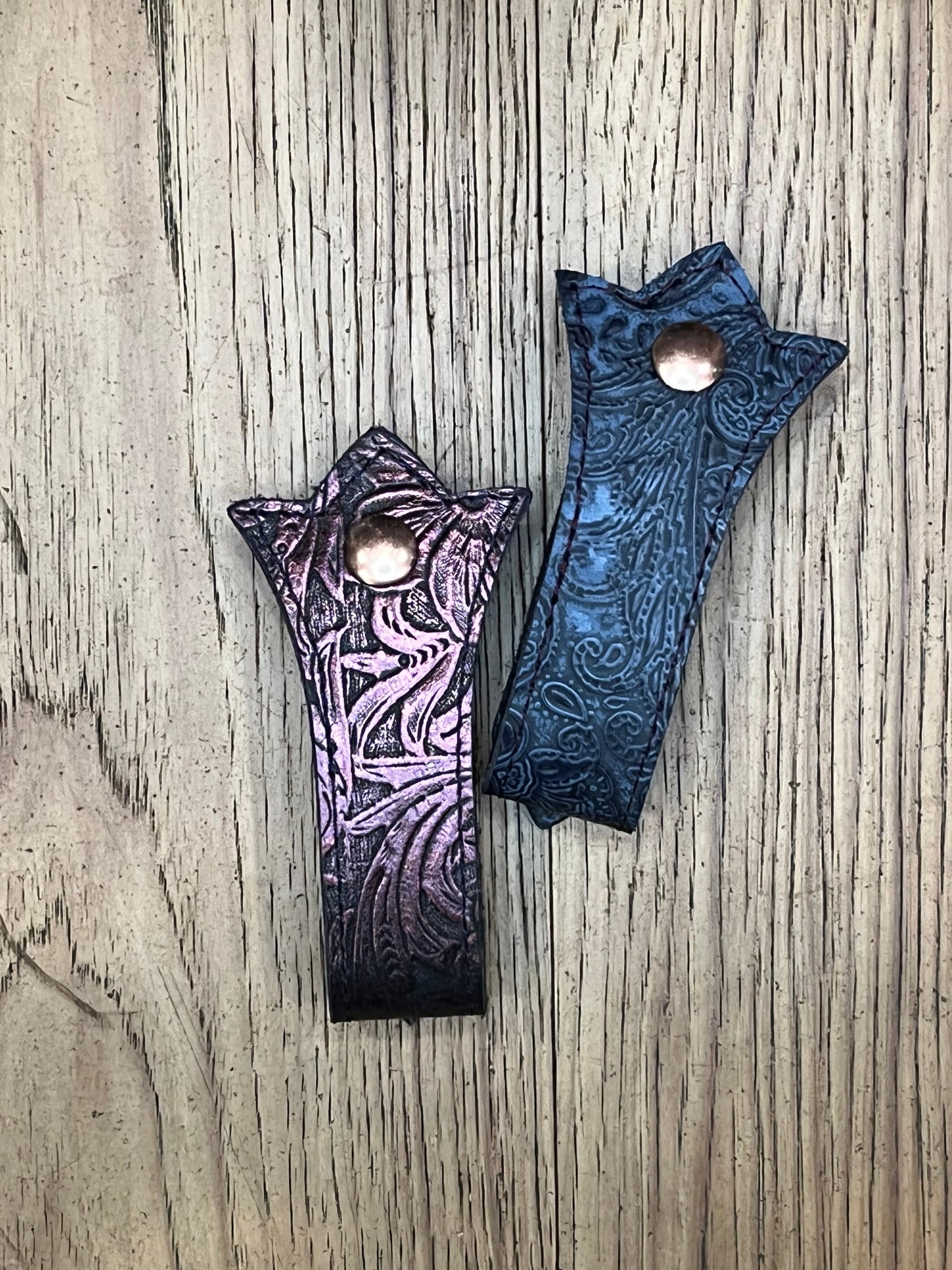 Two Schmancy double layered and stitched mug frogs in the Cordelia colorway. One is a purple floral leather with black stitching and a rose gold snap, the other is a blue paisley leather with black stitching and a rose gold snap. 
