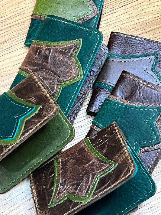 Stack of fiddlehead fern wallets. Variants include colors of dark forest green, apple green, brown and brown floral textured leather with either green or brown stitching. 