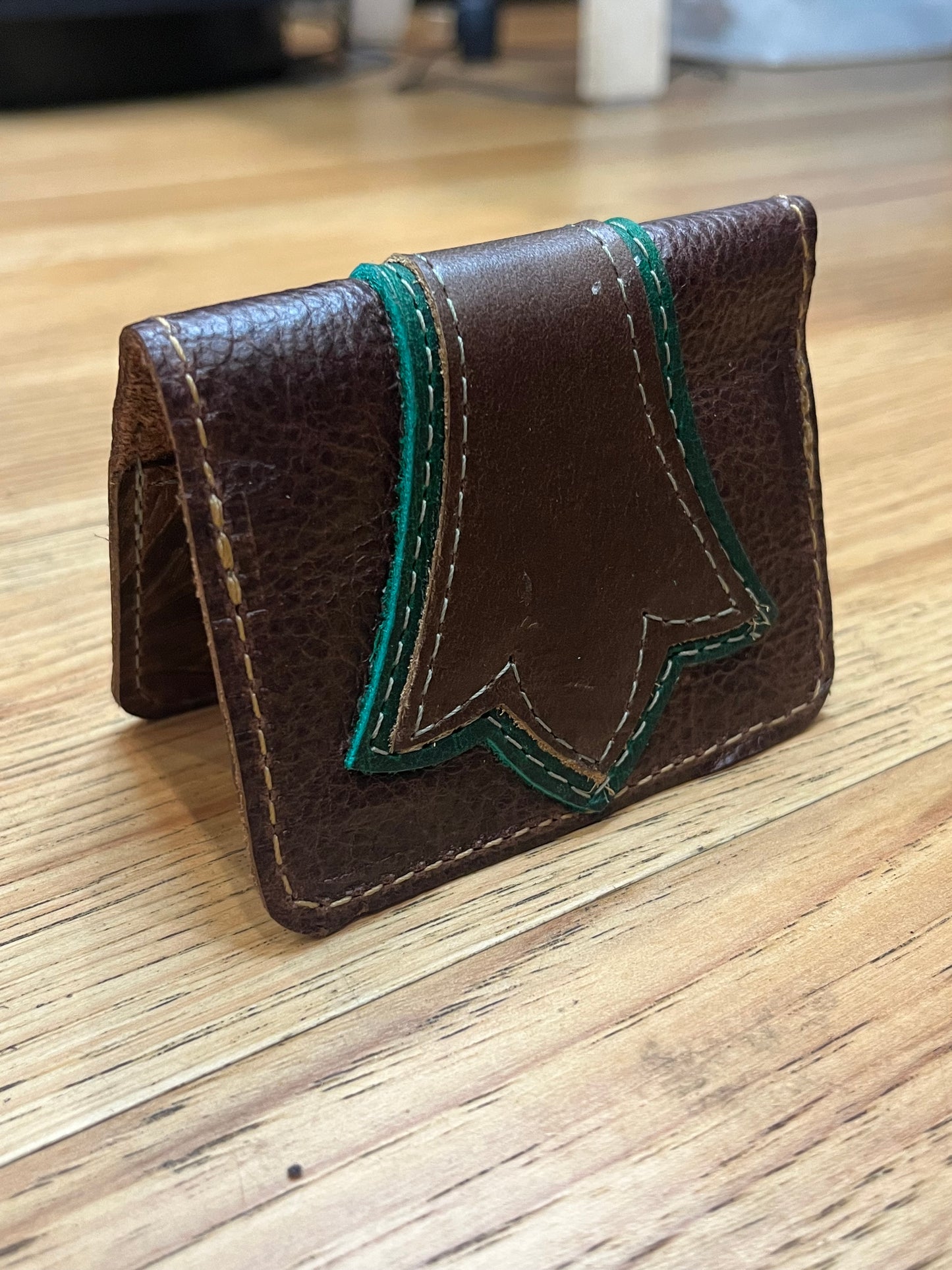 A wallet folded in half showing a base color of brown leather with a layered trident accent in dark green and brown leather with green and brown stitching. 