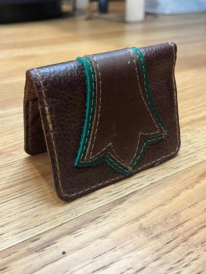 A wallet folded in half showing a base color of brown leather with a layered trident accent in dark green and brown leather with green and brown stitching. 