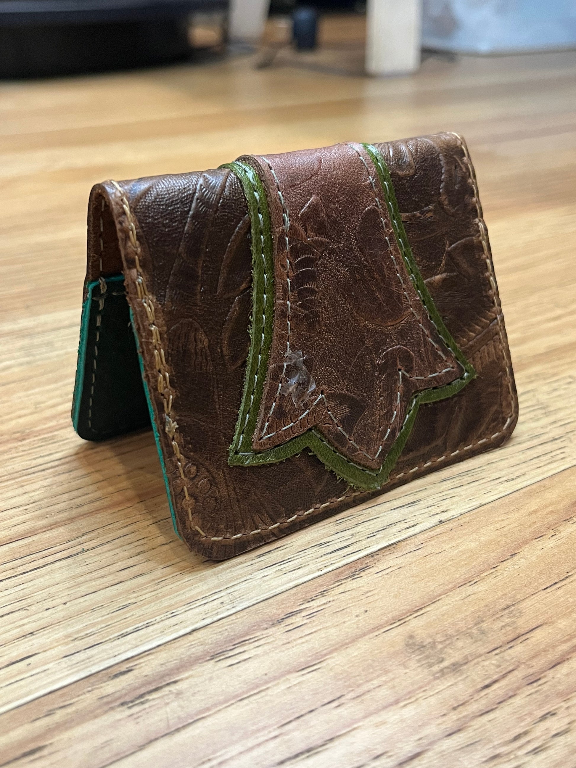 A wallet folded in half showing a base color of brown floral leather with a layered trident accent in light green and brown floral leather with green and brown stitching. 