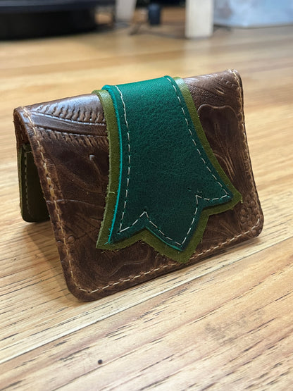 A wallet folded in half showing a base color of brown floral leather with a layered trident accent in dark green and light green leather with green and brown stitching. 
