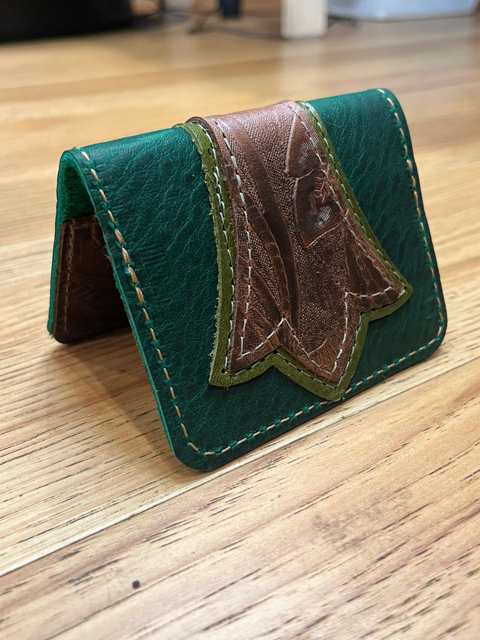 A wallet folded in half showing a base color of dark green leather with a layered trident accent in light green and brown floral leather with green and brown stitching. 
