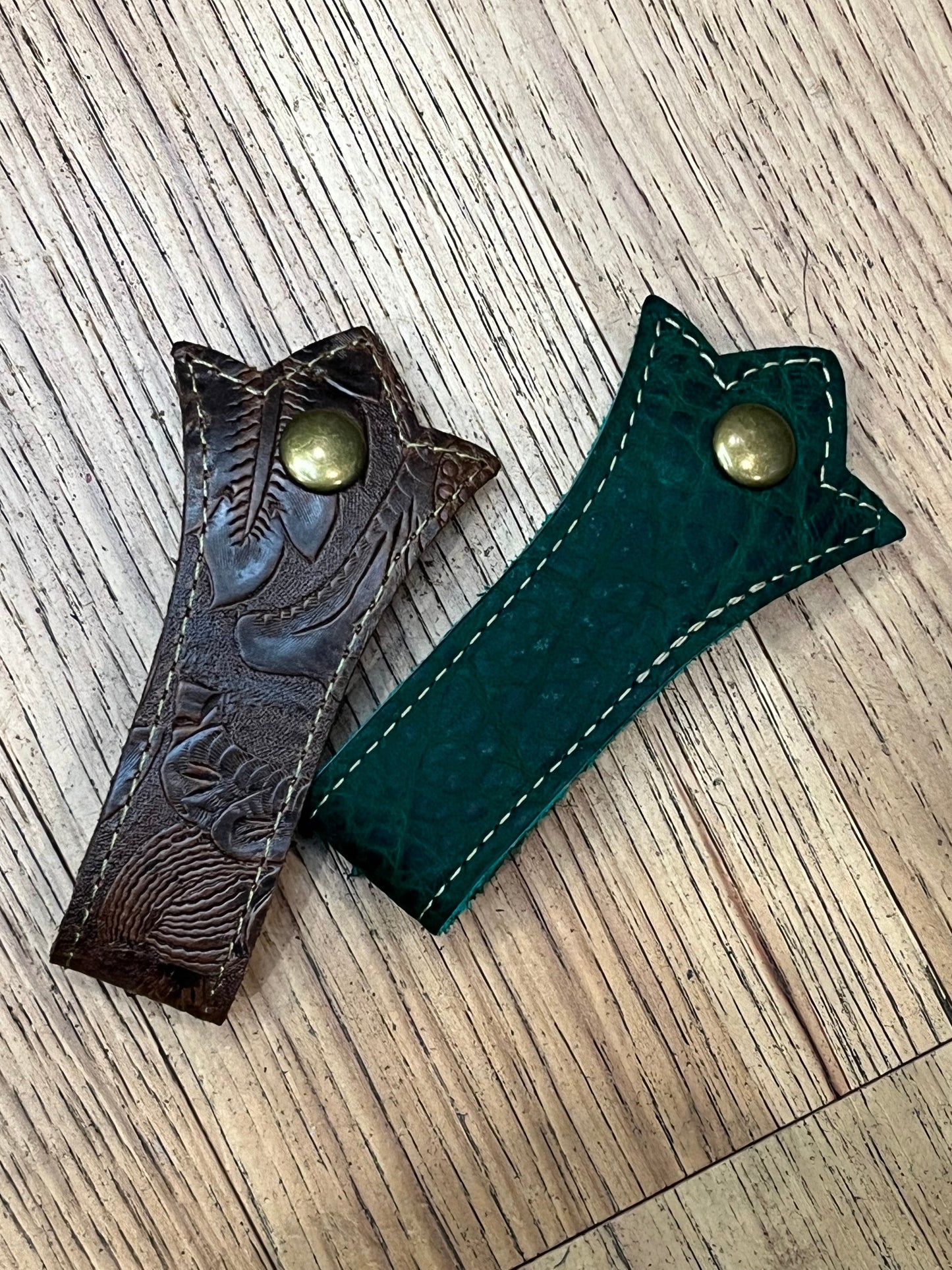 Two Schmancy double layered and stitched mug frogs in the Fiddlehead Fern colorway. One is a brown tooled  leather with tan stitching and a brass snap, the other is a dark green leather with tan stitching and a brass snap. 