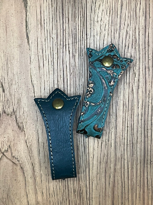 Two Schmancy double layered and stitched mug frogs in the tortuga colorway. One is a teal floral leather with bronze stitching and a brass snap, the other is a teal leather with bronze stitching and a brass snap. 