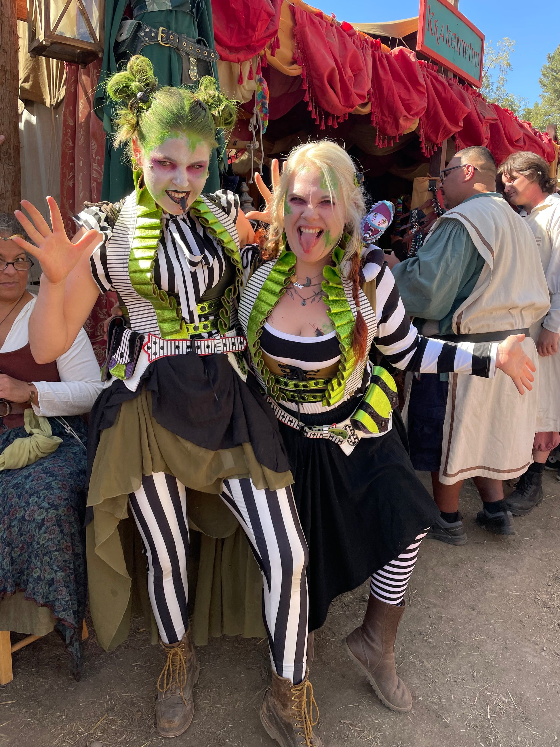 two femme individuals in Beetejuice inspired costumes smiling and sneering at the viewer. each is waering a Showtime Ruff and Showtime Belt. The ruffs are black and white striped leather with lime green leather ruffs. The belts are black and white striped with black stingray, metallic purple, metallic red, and metallic lime green leather accents.