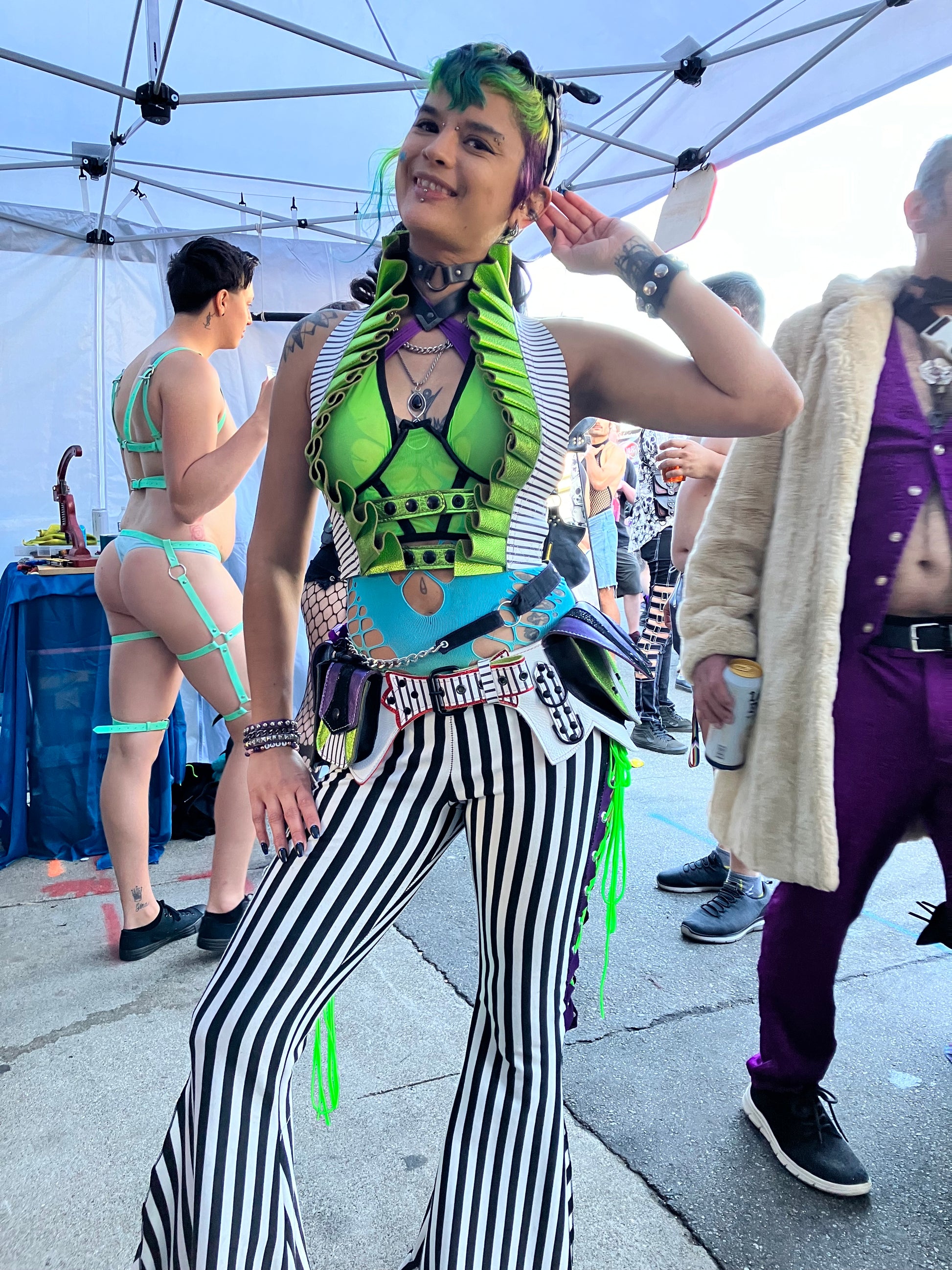 Femme individual wearing black and white striped pants and a lime green top, showing a Showtime Ruff Top in striped leather with a metallic lime leather ruff and a Showtime Regal Belt in the same leathers with purple and red leather accents. Individual is standing, smiling at the viewer with on hand up touching their ear. 