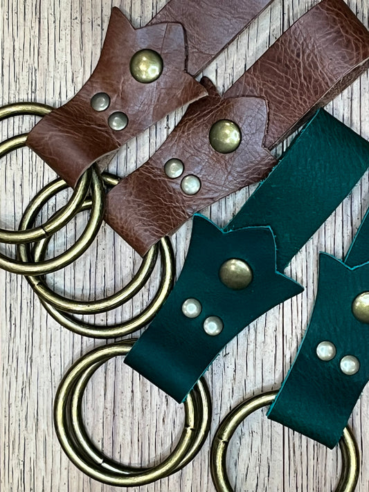 Two pairs of skirt hikes, one made with brown leather in a trident shape with brass snaps and rings and the other made in dark green leather with brass snaps. 
