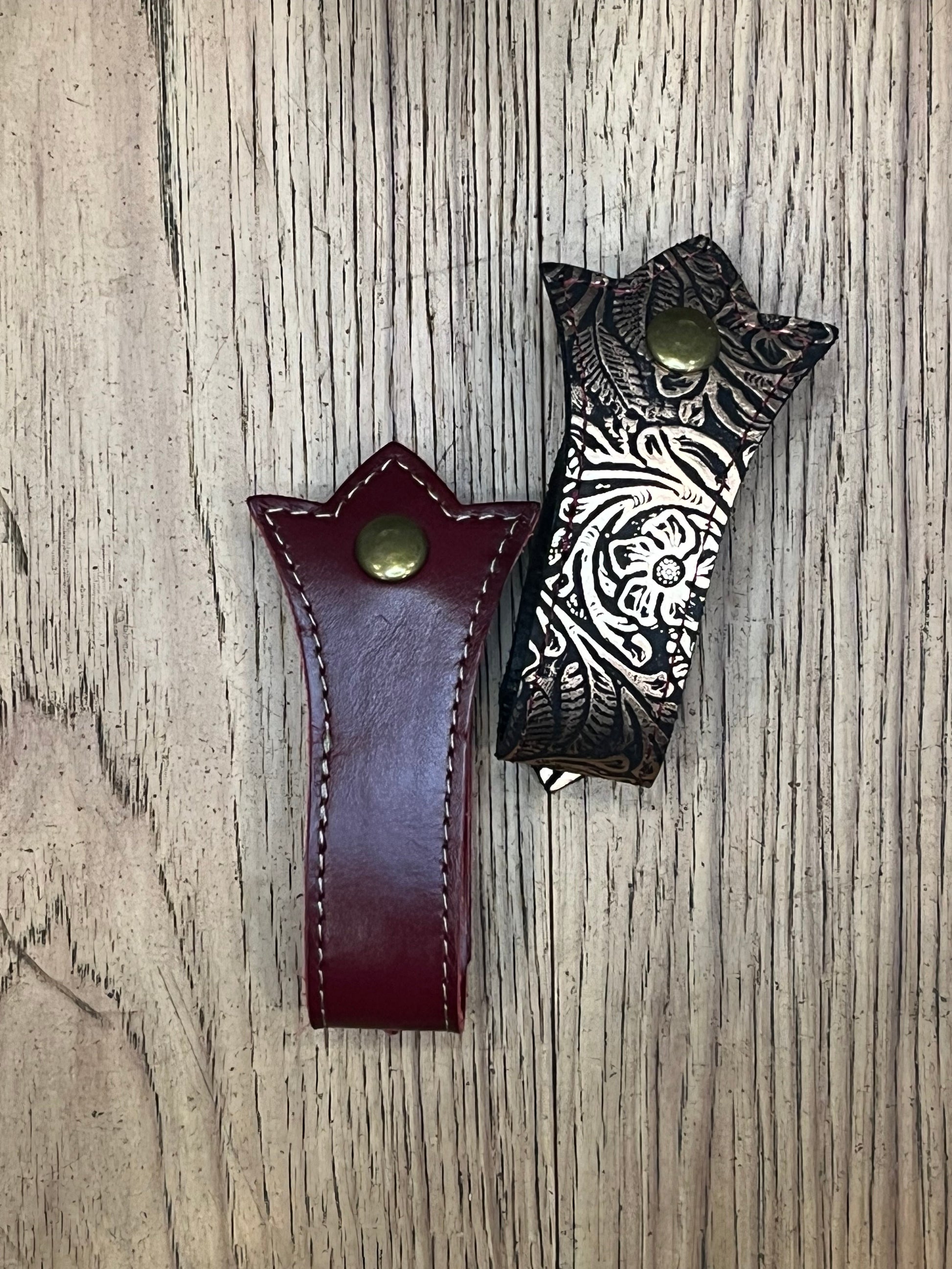 Two mug frogs in a trident shape. One is bronze floral leather with red stitching and the other is dark ed leather with bronze stitching. 