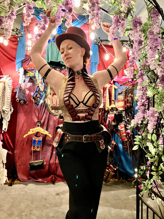 Individual wearing rose rouge ruff top, black pants, rose rouge regal belt and a top hat stands in front of the booth with both hands grasping the archway covered in wisteria and looks at the viewer with a saucy expression 