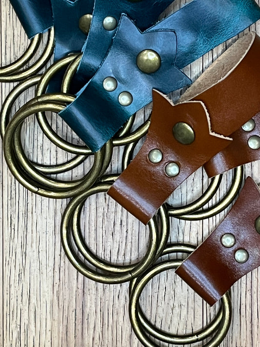 Two pairs of skirt hikes made in a trident shape. One made with teal leather, brass snaps and rings and the other made in brown leather with brass snaps and rings. 
